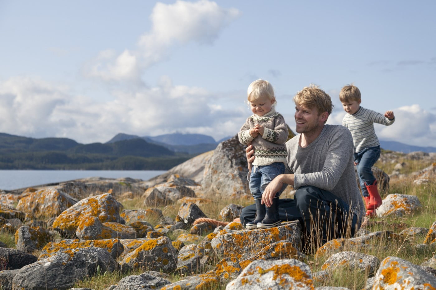 Far og to små born nyter ein dag ute i naturen, sit på steinar dekka av lyng på ein solfylt dag.