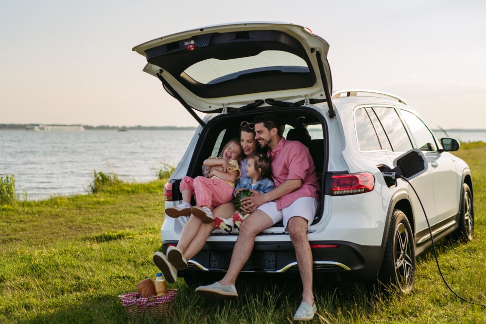 Familie på fire koser seg i bagasjerommet på bilen, parkert ved ein innsjø med ein piknikkurv ved sida av.