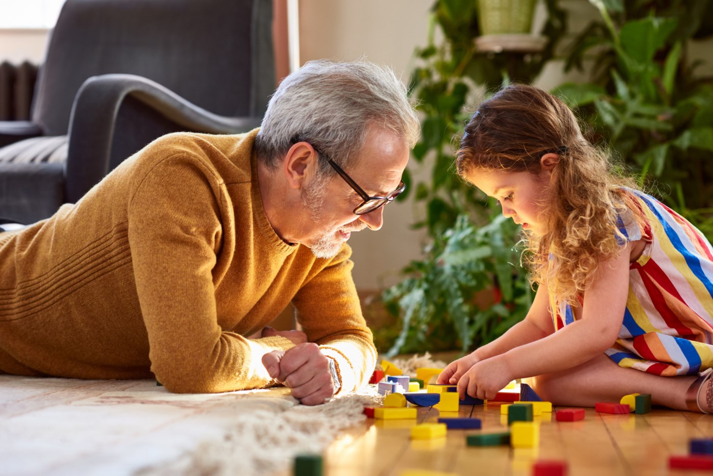 Ein eldre mann med grått hår og briller byggar Lego på golvet saman med sitt barnebarn med krøllete hår i ein stripete kjole.