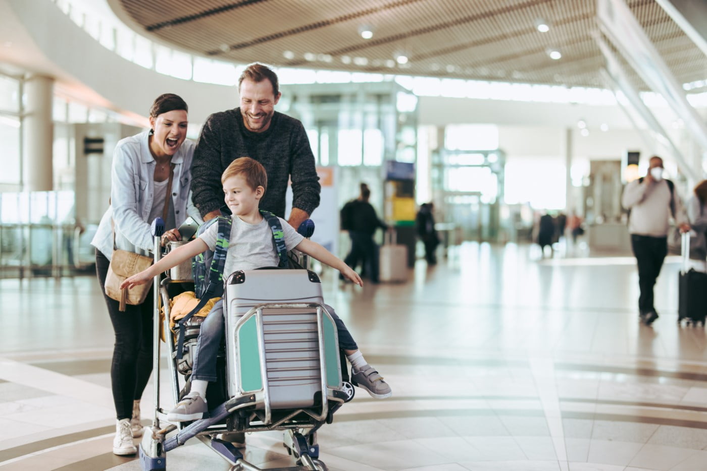 Familie på flyplassen ler og har det gøy, med eit barn som sit på ein bagasjetralle.