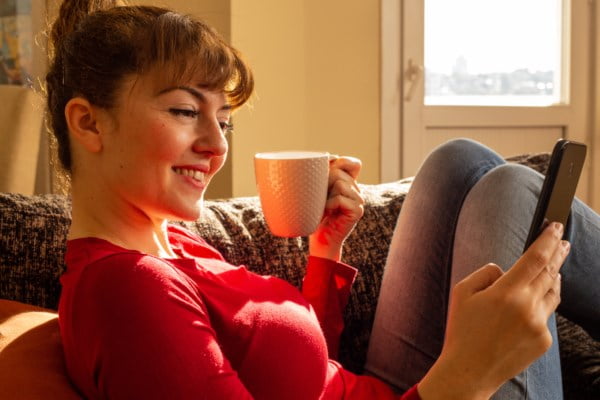 Kvinne i sofa med mobil og kaffikopp i vårlys – får oversikt over økonomien med mobilen og eit roleg smil.