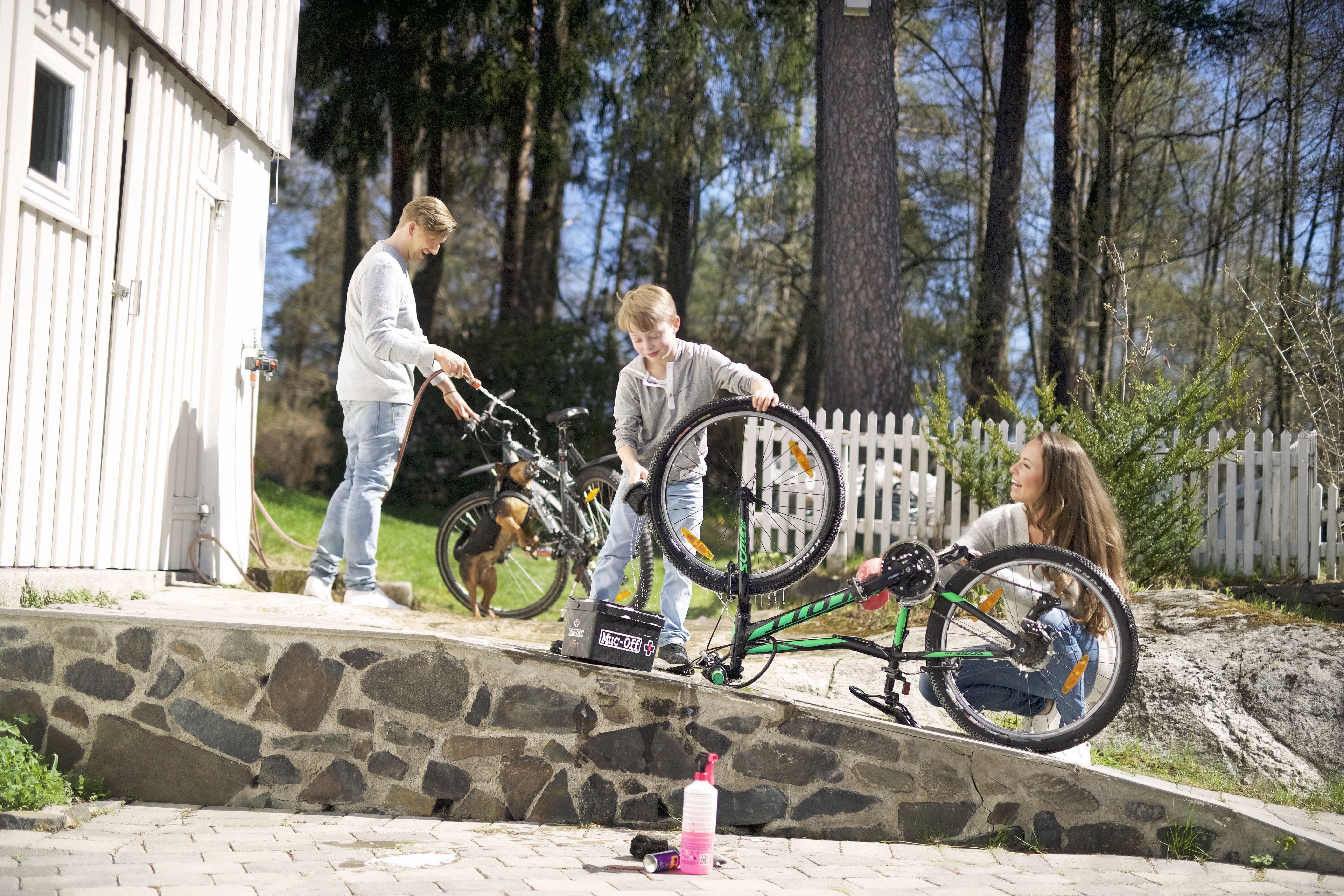 Tre personar og ein hund ute i hagen reparerer syklar ved ein steinmur, med verktøykasse og drikkeflaske på bakken.
