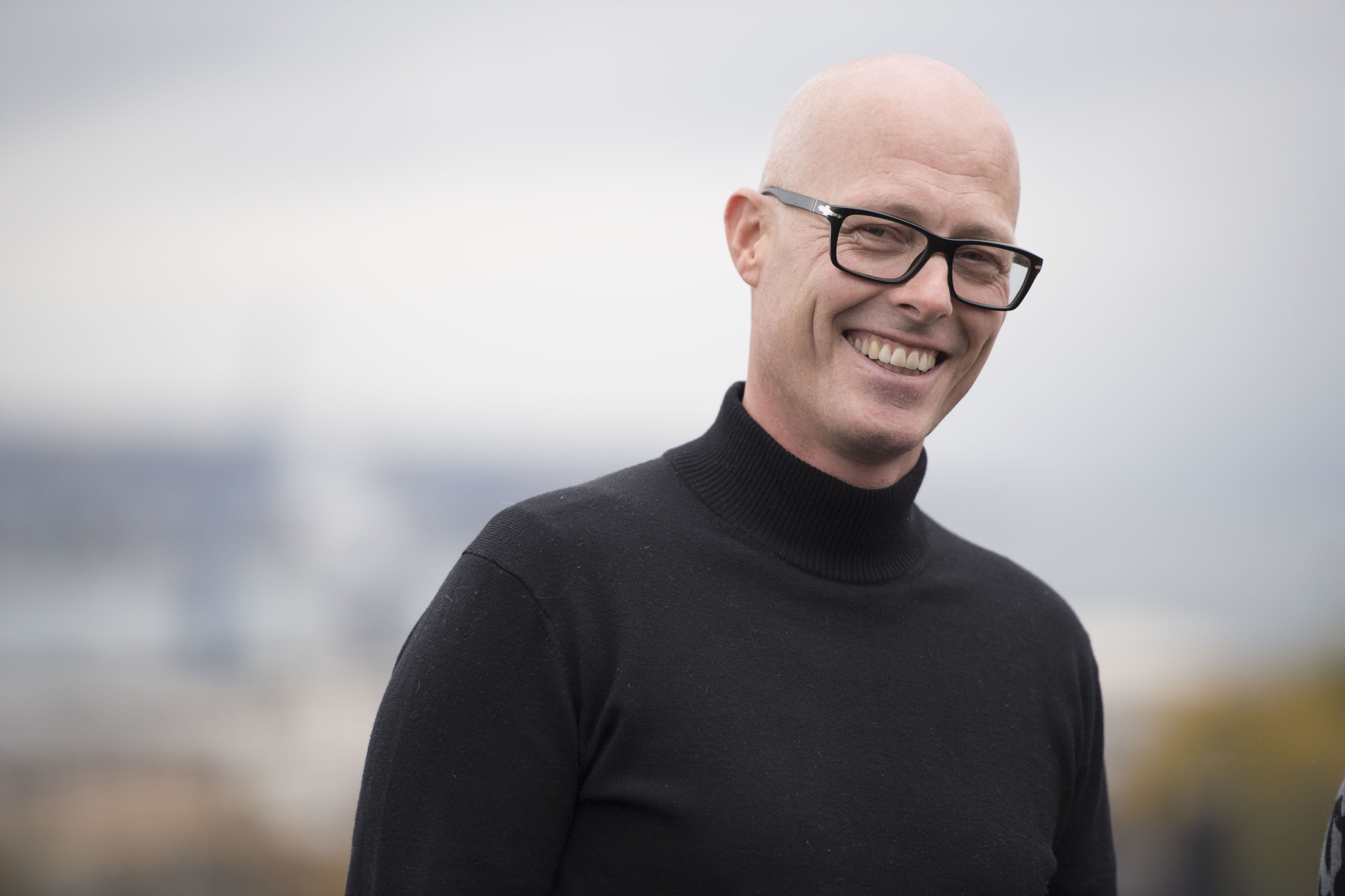 Person med svart høghalsa genser står ute med uklar bybakgrunn.