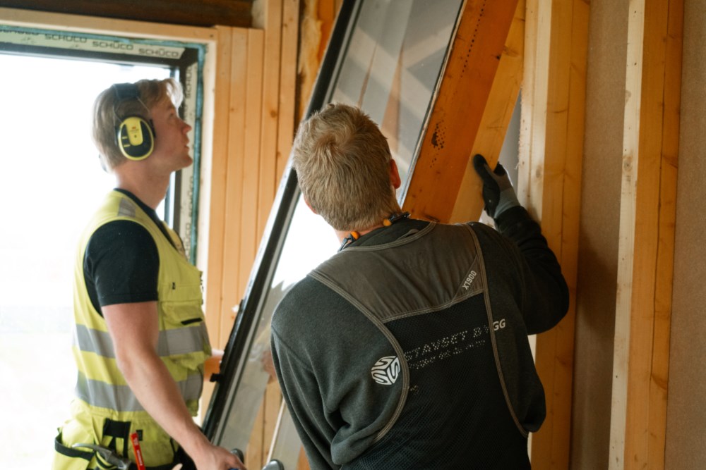 To personar monterer eit stort vindauge i ei trekonstruksjon, med fokus på samarbeid og presisjon i arbeidet.