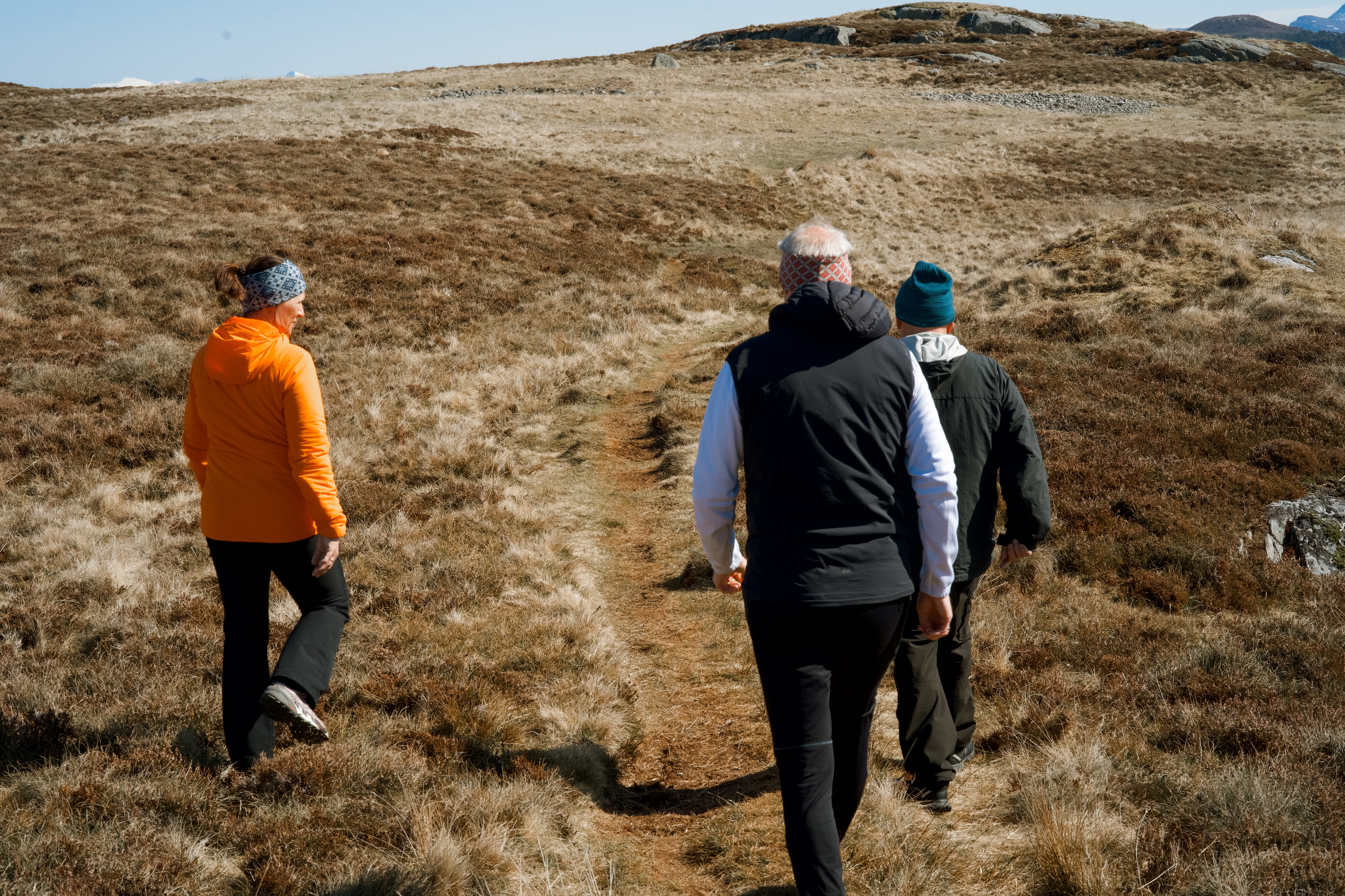 Tre personar går på ein smal sti i eit ope landskap med gras og lyng.