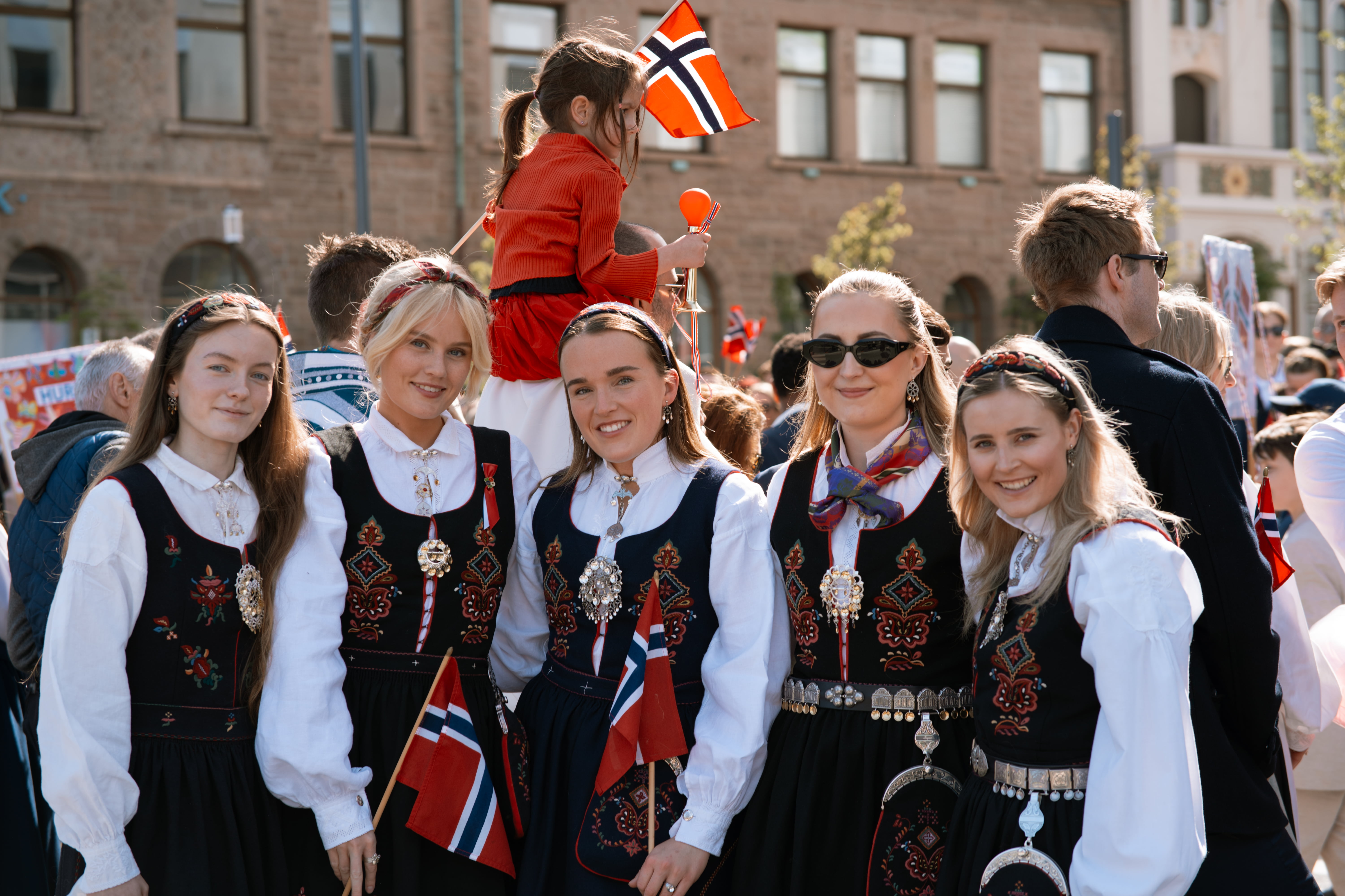 Fem jenter som står ved sida av kvarandre framfor dei som ser på 17. mai-toget.