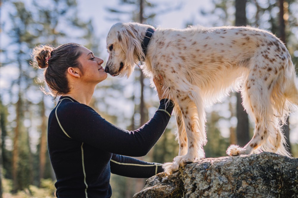 Kvinne i svart genser held ein kvit og brunflekkete hund som står på ein stor stein i skogen, med tre og blå himmel i bakgrunnen.