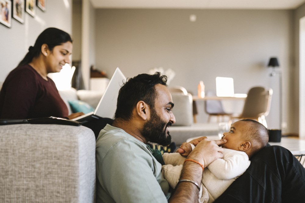 Familie på tre der mor sit i sofaen med PC, medan far sit på golvet og leikar med babyen.