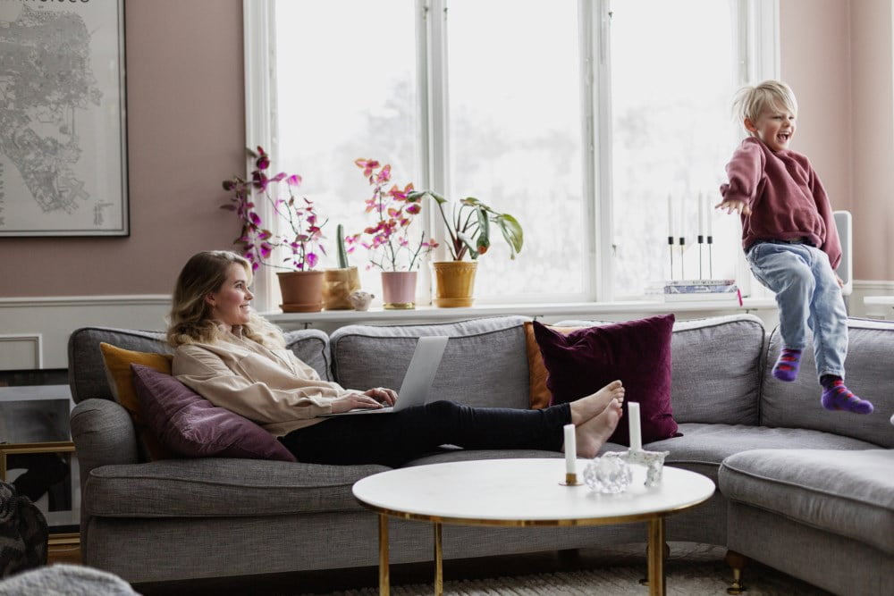 Mor som sit i sofaen og jobbar, medan barnet hoppar ved sidan av.