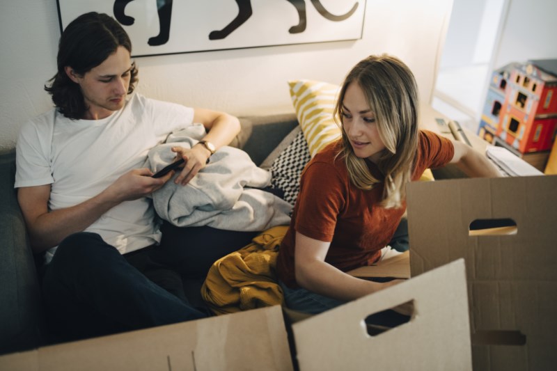 Ung jente og gutt sitter i sofaen blant flytteesker med mobil