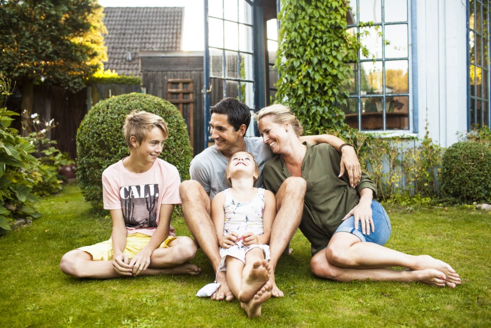Familie med to barn som sit på graset ute i hagen ein sommarkveld.