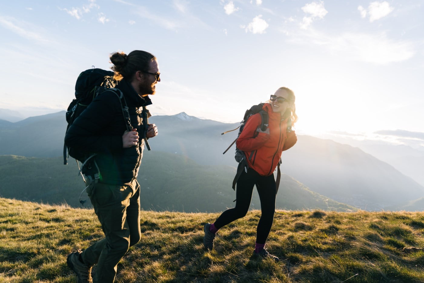 Mann og kvinne med ryggsekkar går på ein fjelltopp med solnedgang i bakgrunnen.