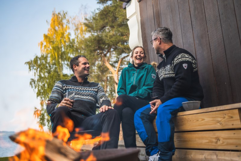 Tre menneske sit i hytteveggen framfor eit bål