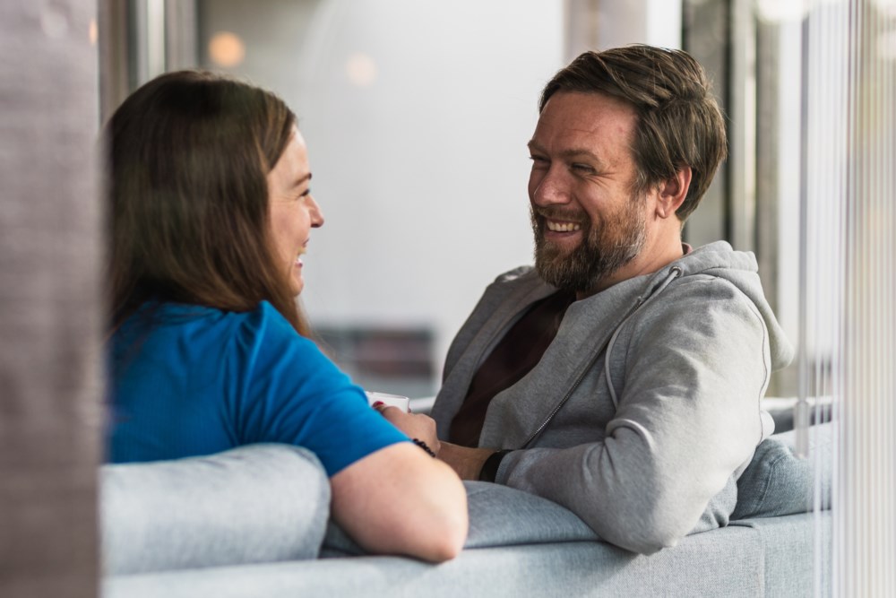 Ser inn gjennom vindauget på kvinne og mann i sofaen som smiler til kvarandre